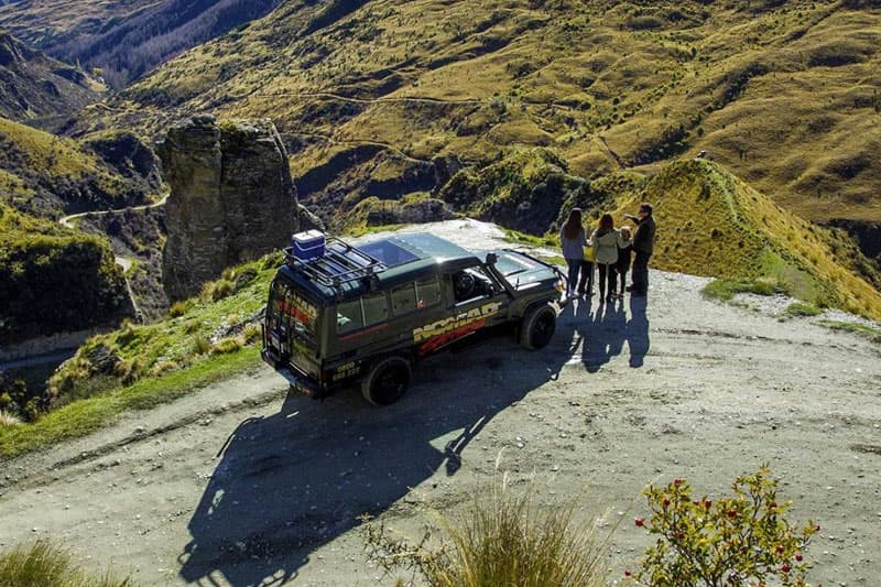 A red "Shotover Jet" boat is captured mid-turn, creating a dramatic spray of water that arches over the boat, partially obscuring the view of the passengers. The intense motion of the boat contrasts with the blurred background of trees and landscape, emph