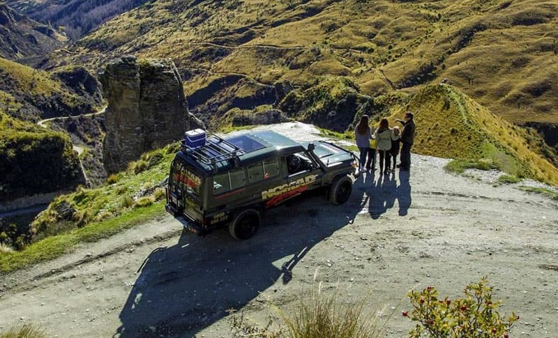 A red "Shotover Jet" boat is captured mid-turn, creating a dramatic spray of water that arches over the boat, partially obscuring the view of the passengers. The intense motion of the boat contrasts with the blurred background of trees and landscape, emph