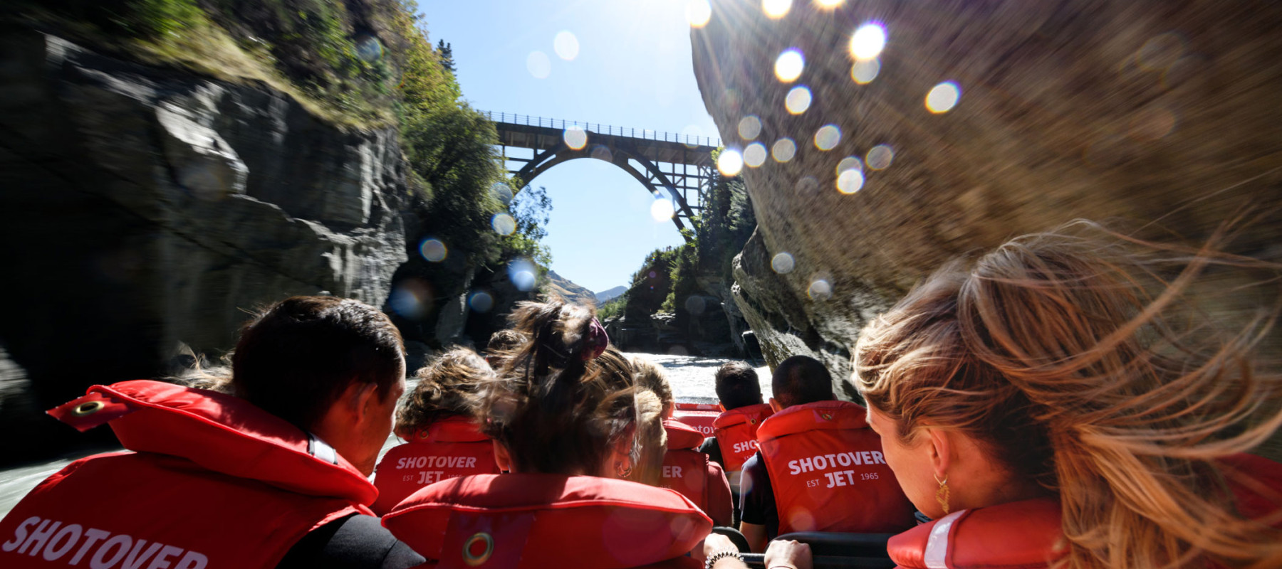 A group of people wearing red "Shotover Jet" life jackets are seated in a jet boat, heading towards a bridge that spans a narrow canyon. The photo is taken from behind the passengers, capturing the su