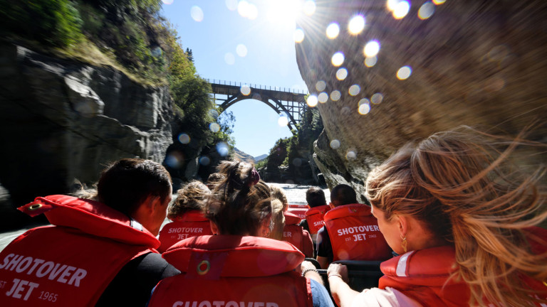 A group of people wearing red "Shotover Jet" life jackets are seated in a jet boat, heading towards a bridge that spans a narrow canyon. The photo is taken from behind the passengers, capturing the su