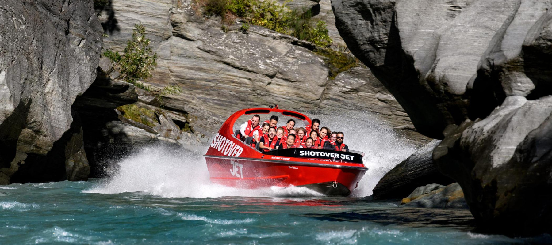 The Shotover Jet boat speeds through a narrow canyon, creating a large splash as it navigates the turquoise waters between towering rocky cliffs. The boat is filled with passengers wearing life jacket