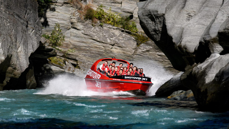 The Shotover Jet boat speeds through a narrow canyon, creating a large splash as it navigates the turquoise waters between towering rocky cliffs. The boat is filled with passengers wearing life jacket
