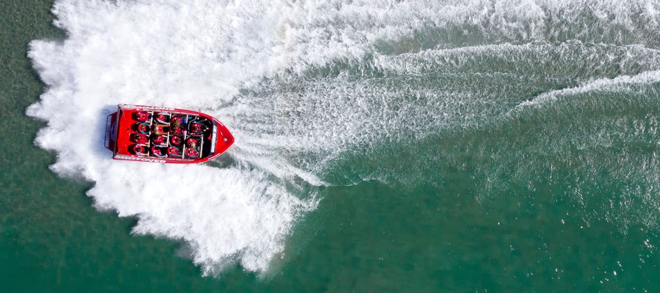 The image shows an aerial view of a red jet boat making a sharp turn on a greenish-blue river, creating wide, sweeping trails of white water behind it, illustrating the excitement and dynamic movement of the high-speed ride