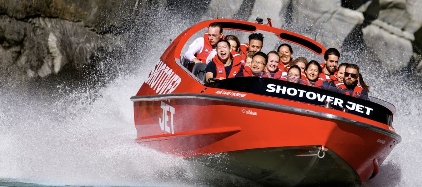 The Shotover Jet speeds through water, creating a large splash as it navigates close to rocky canyon walls. The boat is filled with passengers grinning with excitement, in Queenstown New Zealand