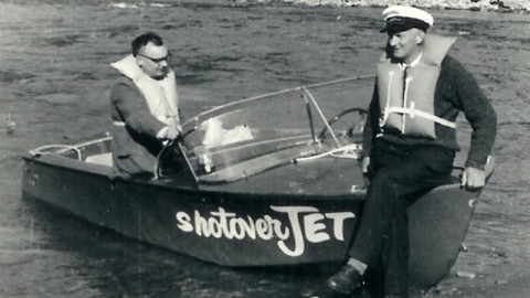 A black-and-white photo of two men in a small "Shotover Jet" boat on a river. One man is seated at the wheel, while the other, wearing a captain's hat and a life jacket, sits casually on the front of