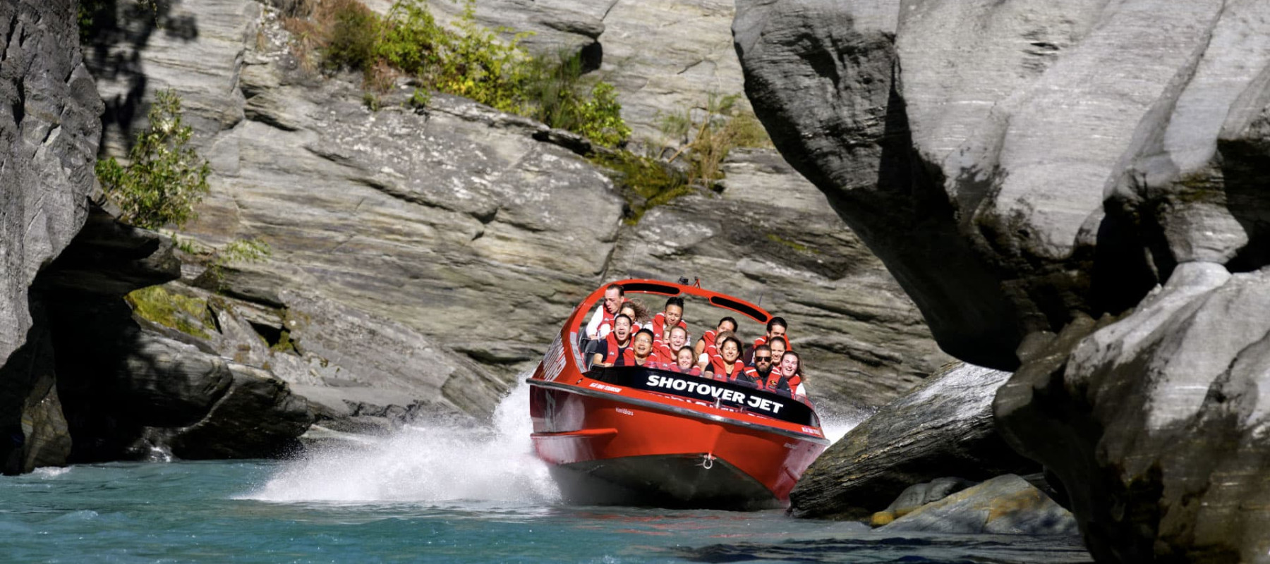 The"Shotover Jet" boat speeds through a narrow, rocky canyon, creating a splash as it maneuvers through the tight space. The boat is filled with passengers wearing life jackets, all smiling and enjoy